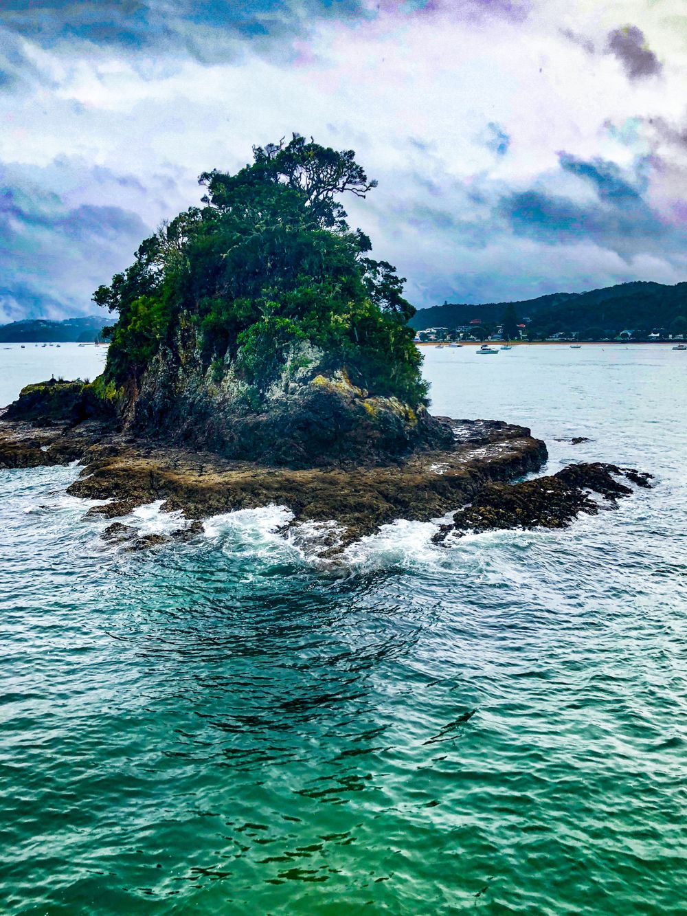Small rocky island with lush green vegetation emerges from turquoise waters under dramatic cloudy skies.