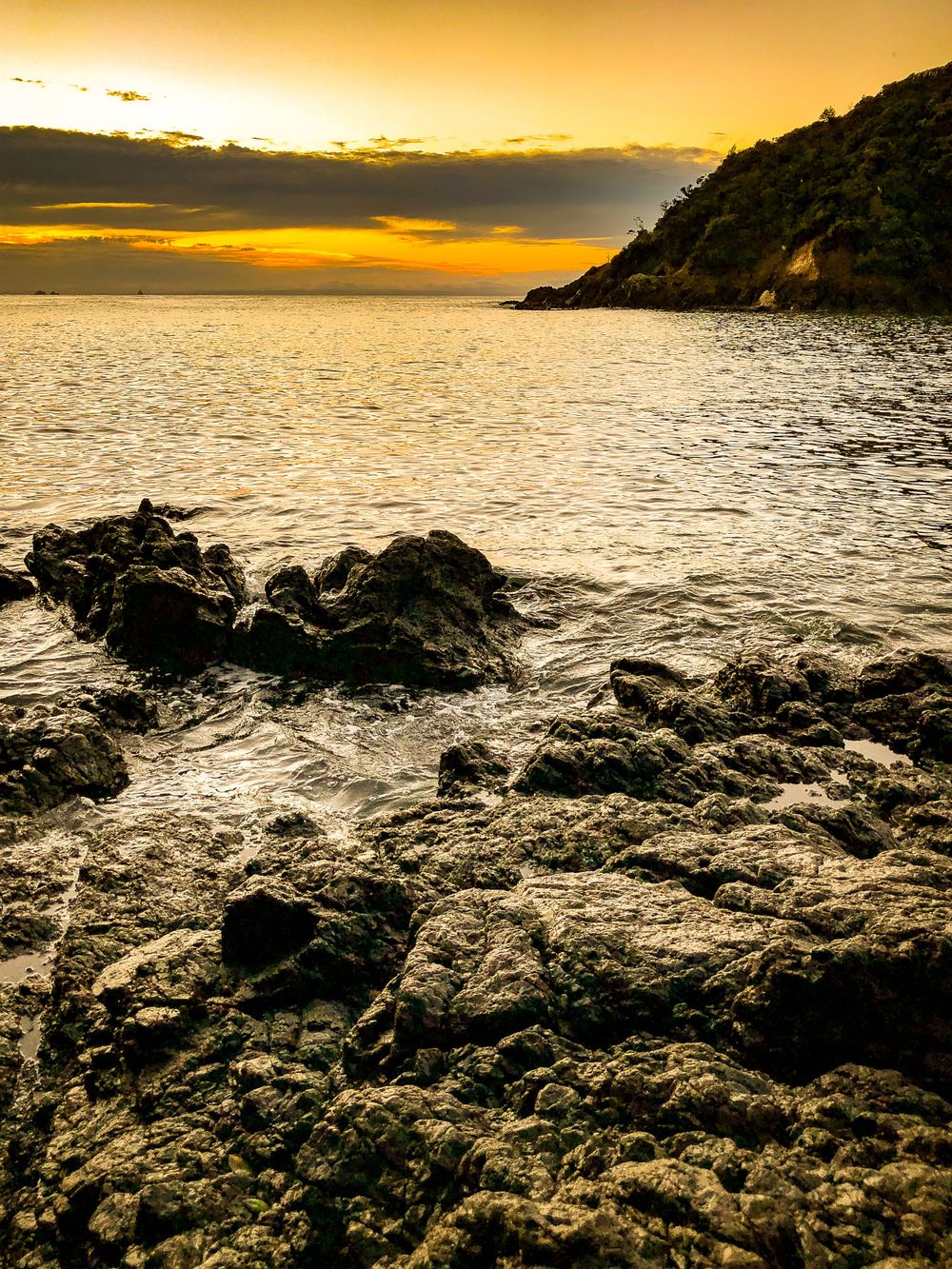 Golden sunset casts warm light over jagged volcanic rocks along the coastal shoreline.