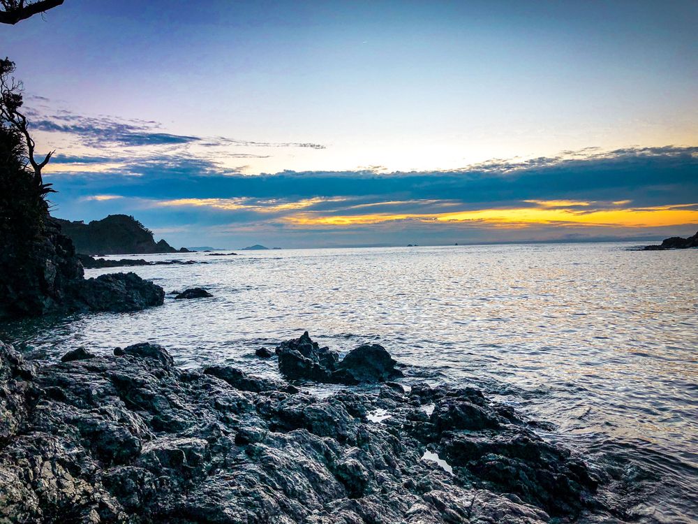 Golden sunset illuminates the horizon over rocky coastal waters with dramatic cloud formations.