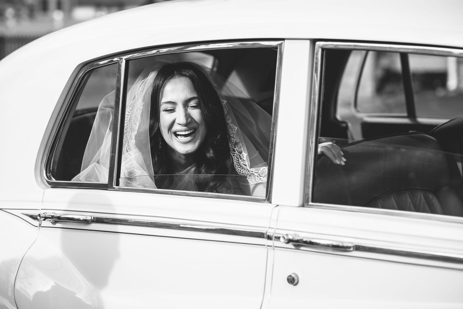 Joyful bride laughing inside a vintage classic white car on her wedding day in black and white.