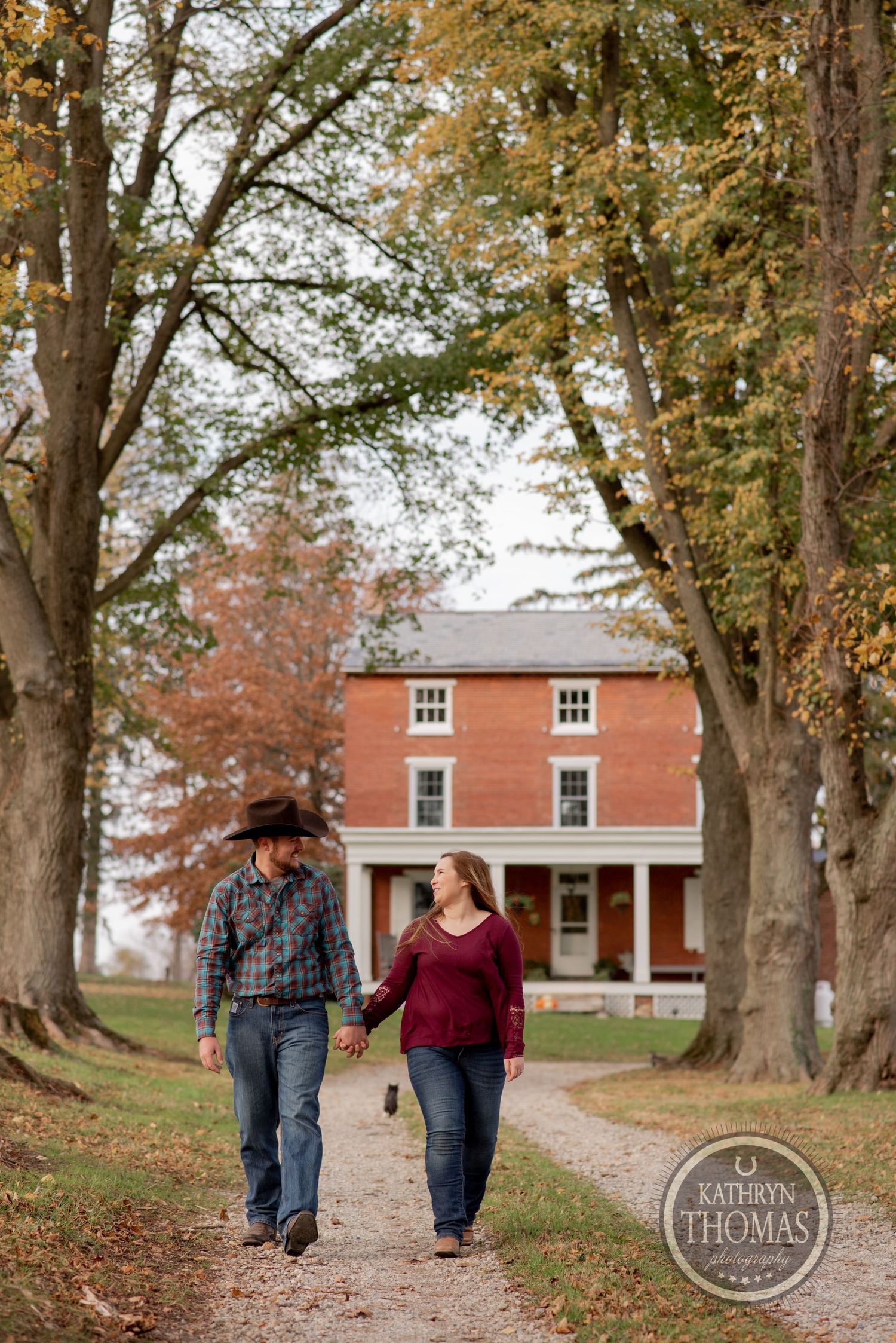 A couple strolls down a tree-lined path in autumn with a brick farmhouse in the background.