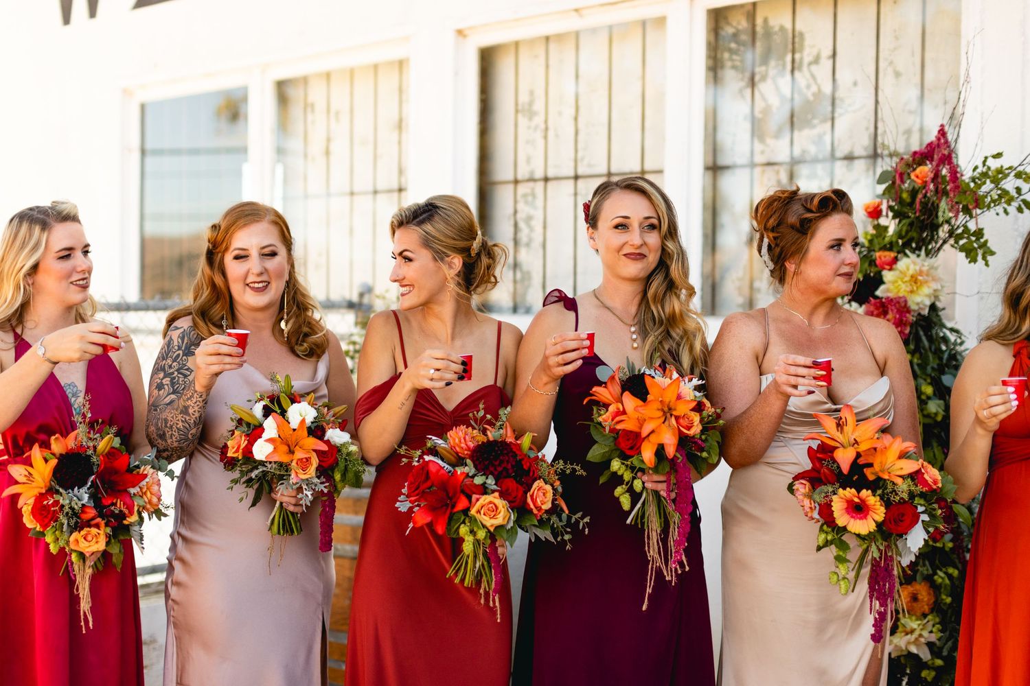 Bridesmaids in mix of red and blush dresses hold autumn bouquets while posing together.