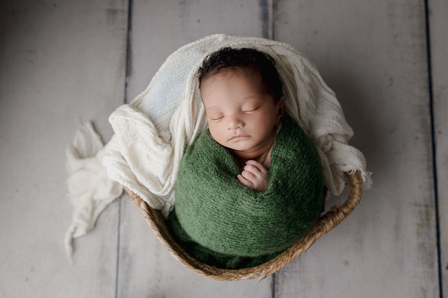 Newborn wrapped in green swaddle sleeping peacefully in wooden bowl with white fabric.