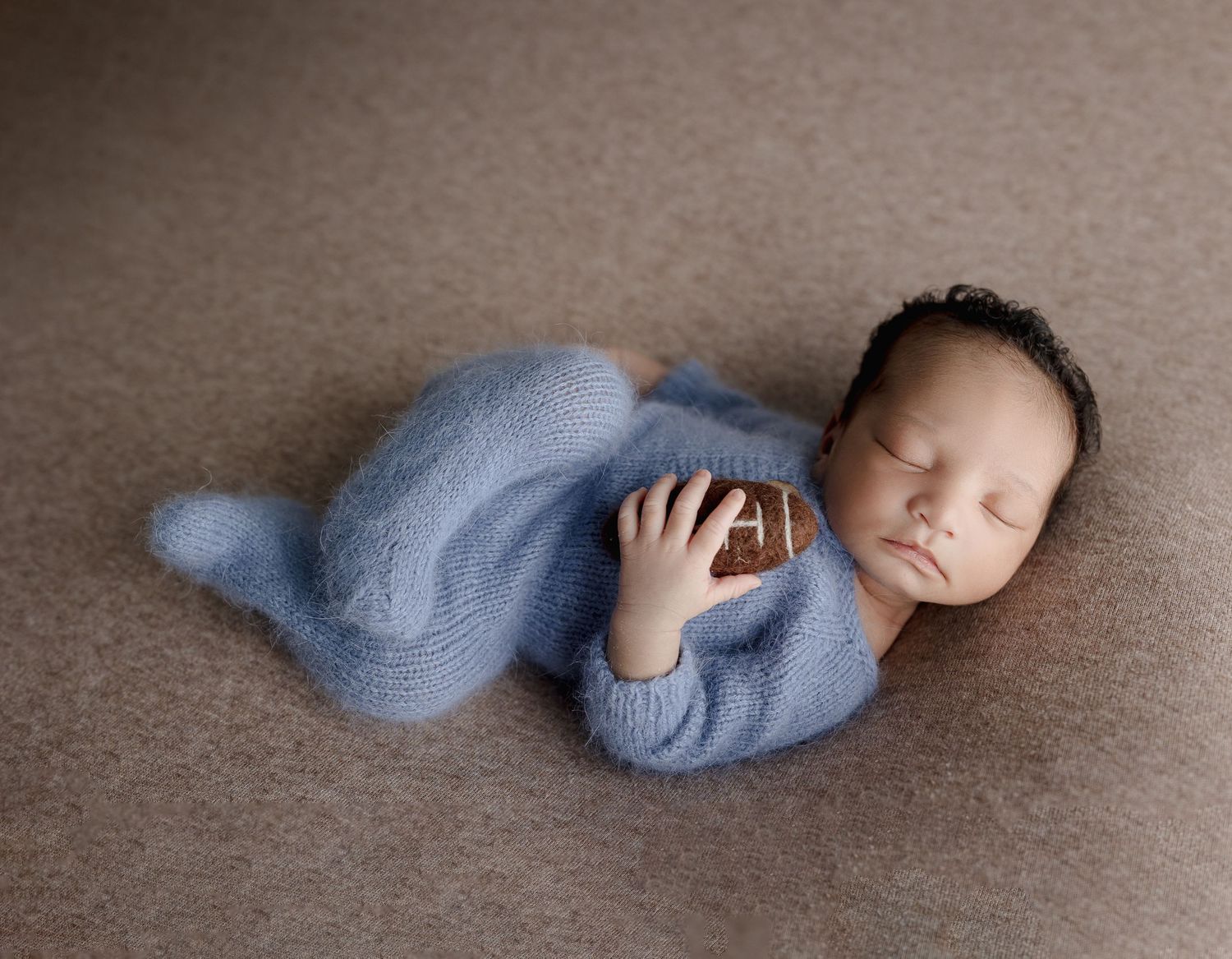 Sleeping baby in light blue knit outfit lying on soft brown backdrop.