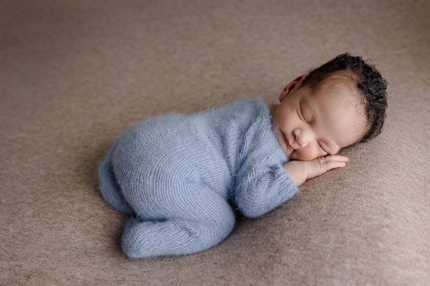 Sleeping newborn in soft blue knit outfit laying on beige backdrop.