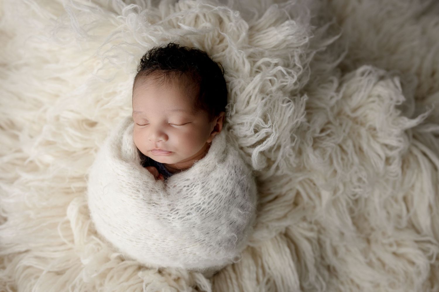 Peaceful newborn wrapped in white swaddle resting on fluffy white fur blanket.