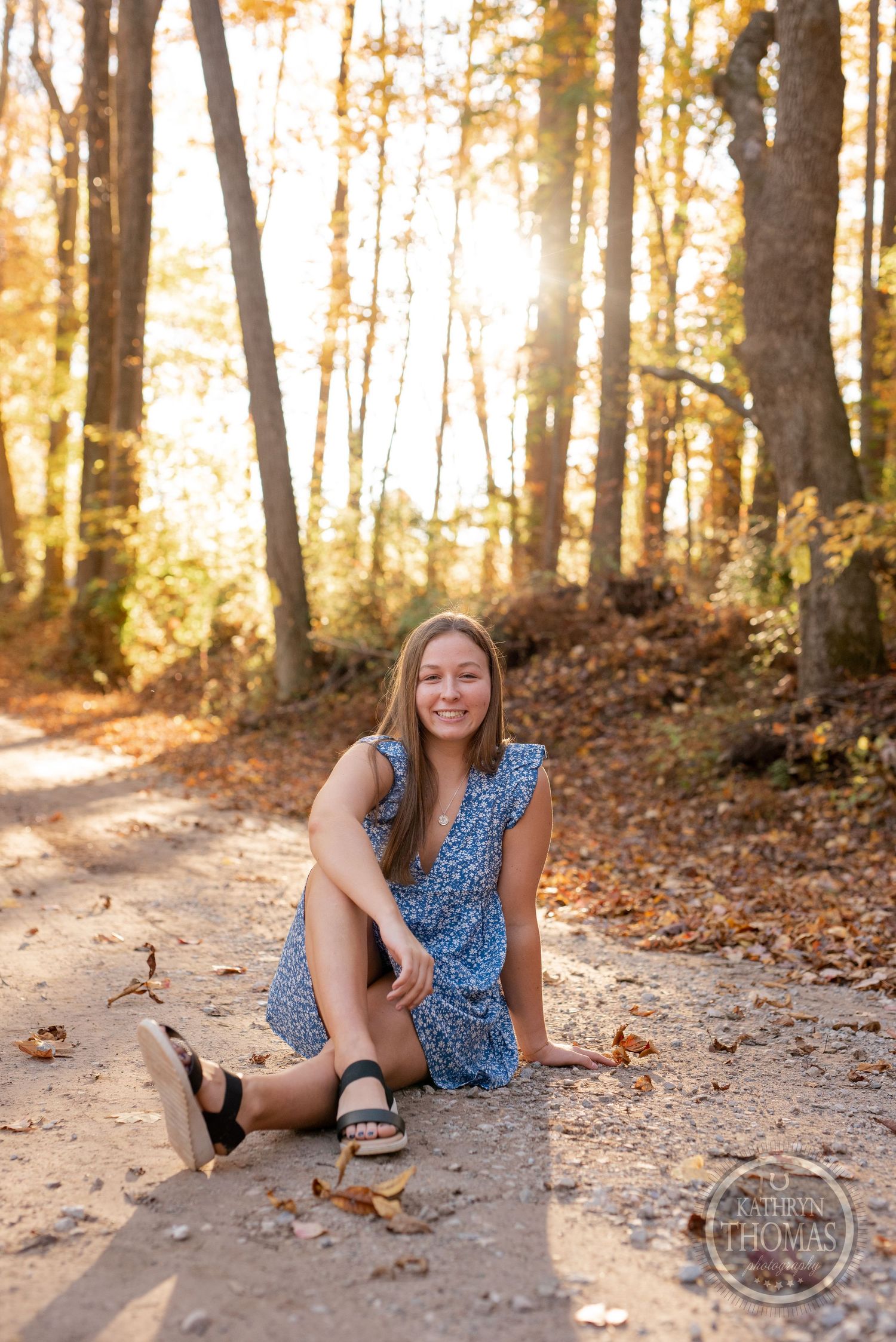 A blue floral dress and sandals on a sunlit autumn forest path with golden leaves scattered around.