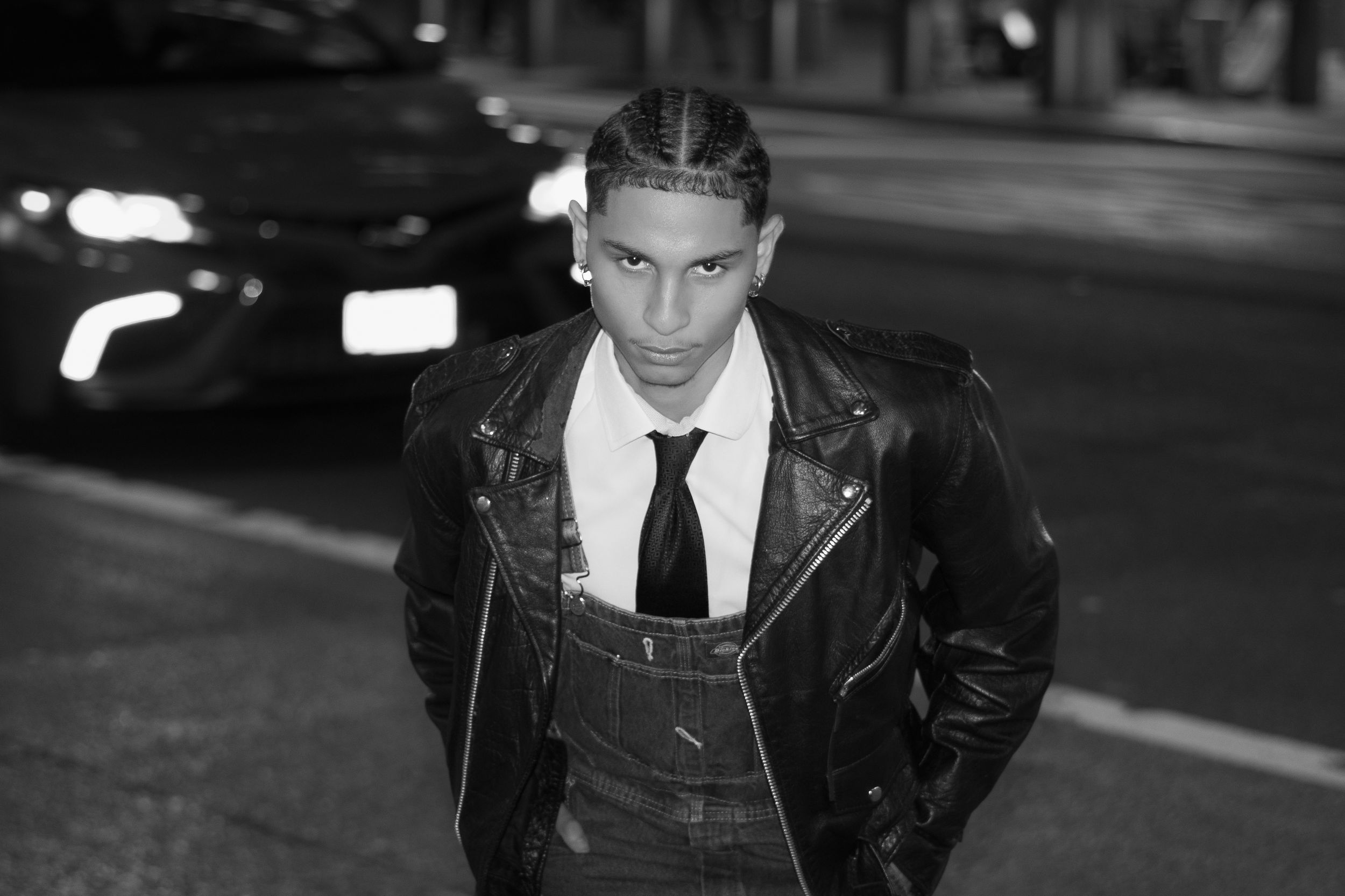 Stylish person in leather jacket and tie poses on nighttime city street with moody lighting.