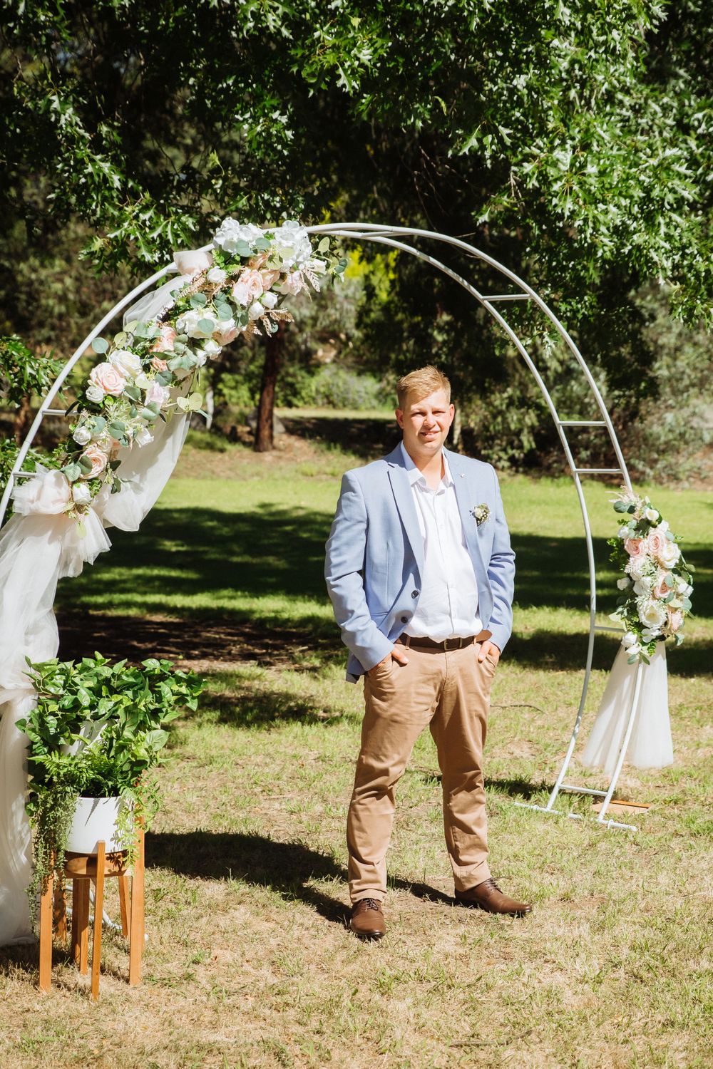 casually dressed blonde groom waiting at arbour decorated in pink adn white for bride