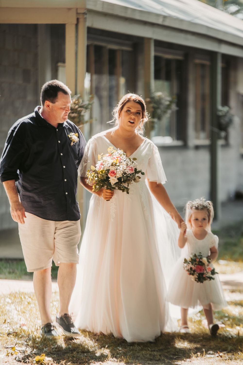 casually dressed father of bride in shorts and dark shirt walking bride down aisle she's holding flower girls hand