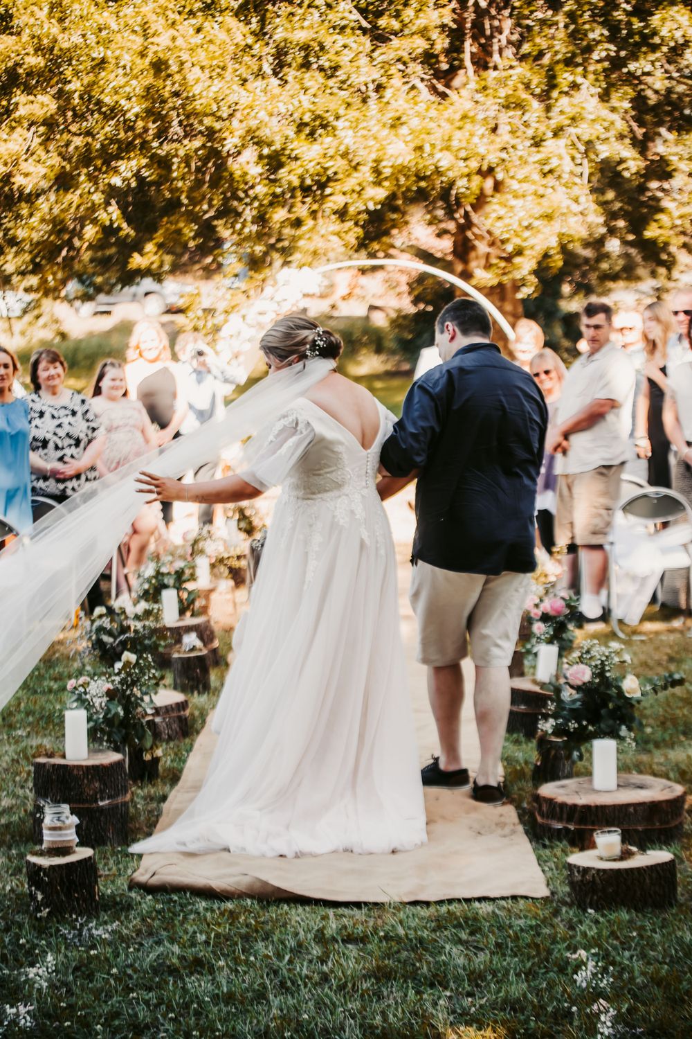 bride in blush dress on father's arm gathers blowing veil in as they walk down aisle