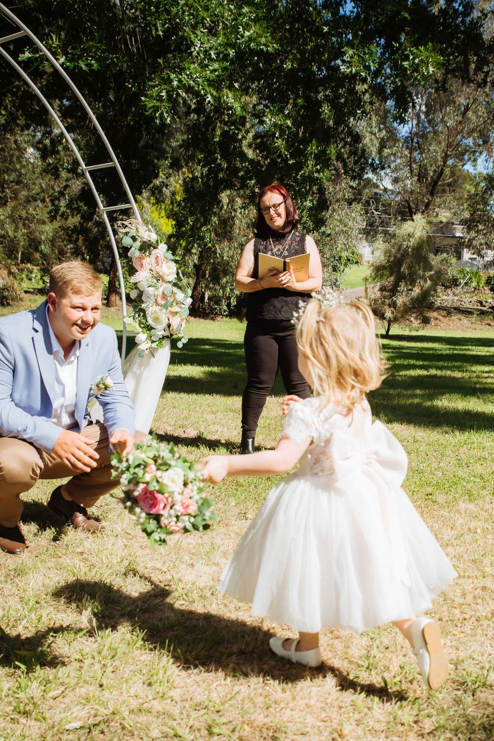 flowergirl in white dress carrying basket of pink and white flowers runs towards groom at outdoor ceremony
