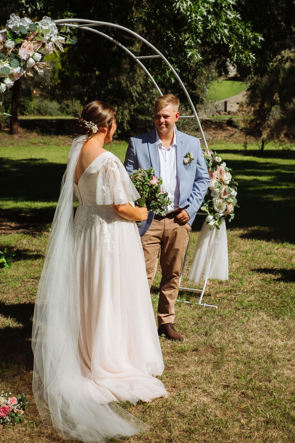 pregnant bride in blush dress and long veil standing at arbour with casually dressed groom