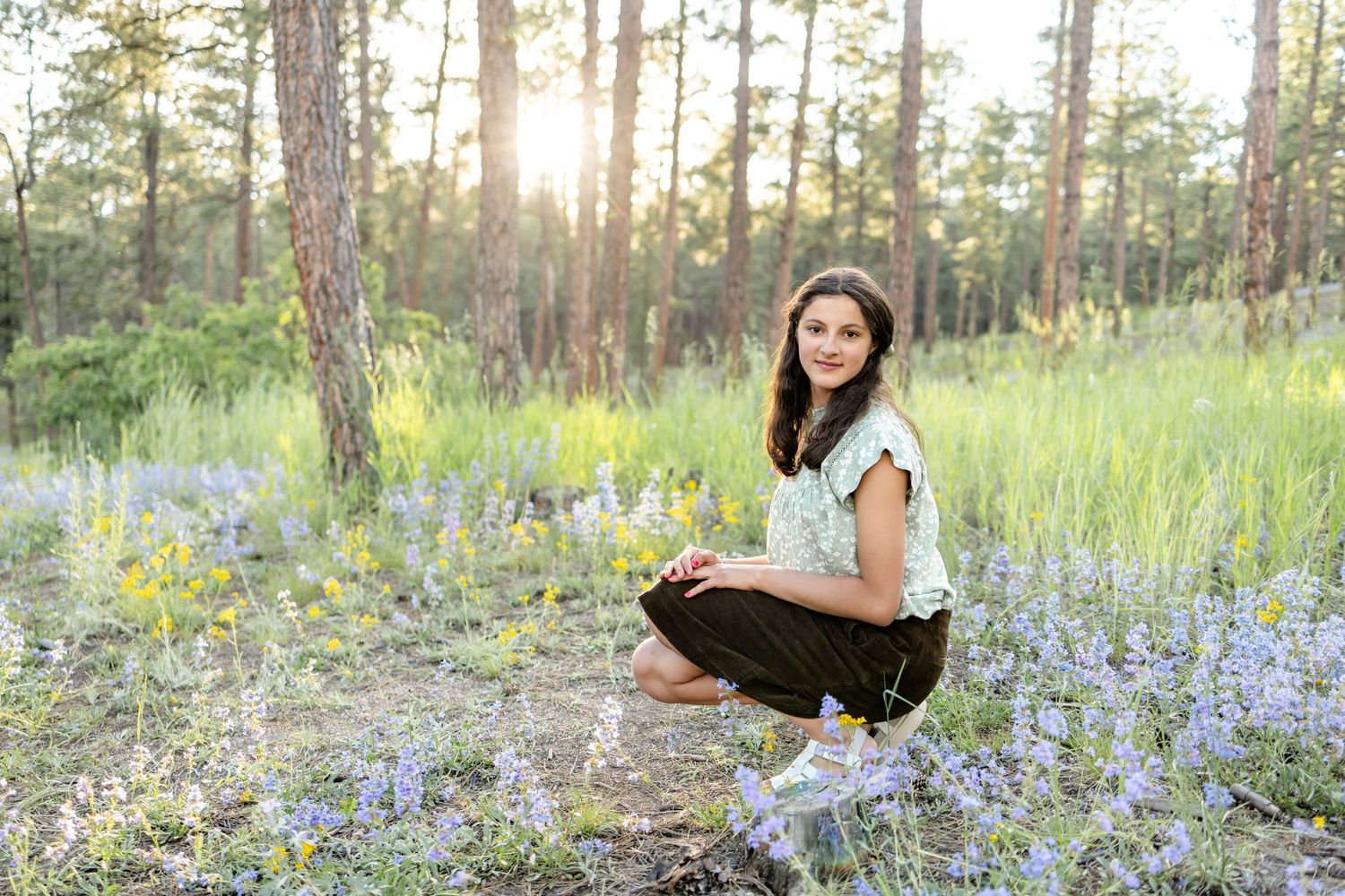 Teen girl squats among the wildflowers at sunset during their family session in Colorado Springs.