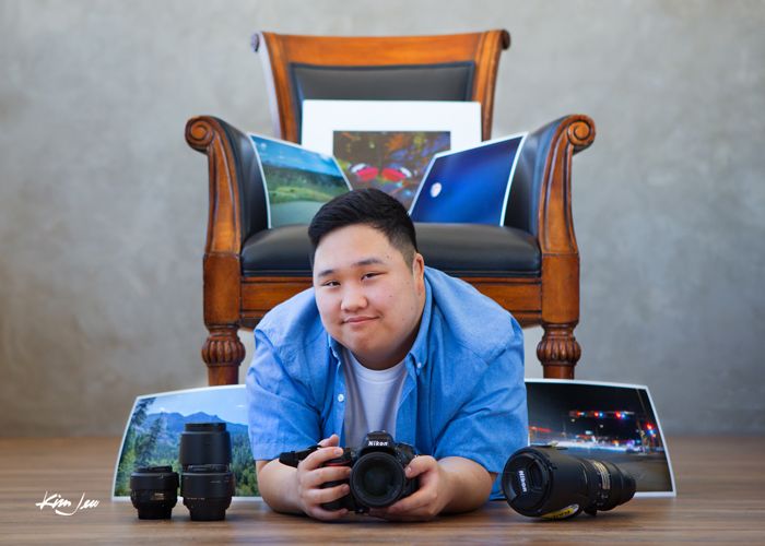 A photographer sits with camera equipment in front of a wooden chair with decorative pillows.