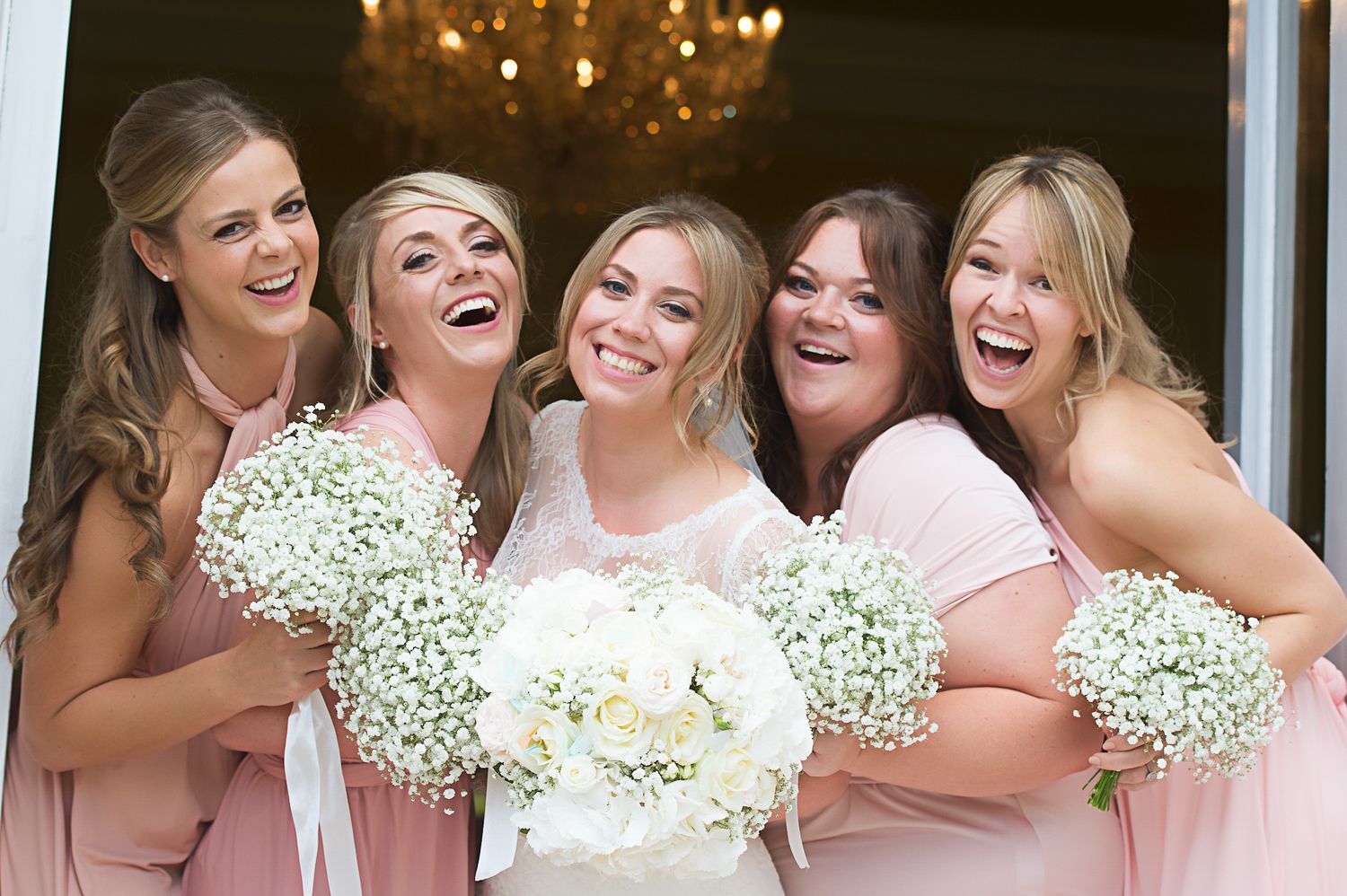 Bridal party with bridesmaids in pink dresses holding baby's breath bouquets share joyful moment.