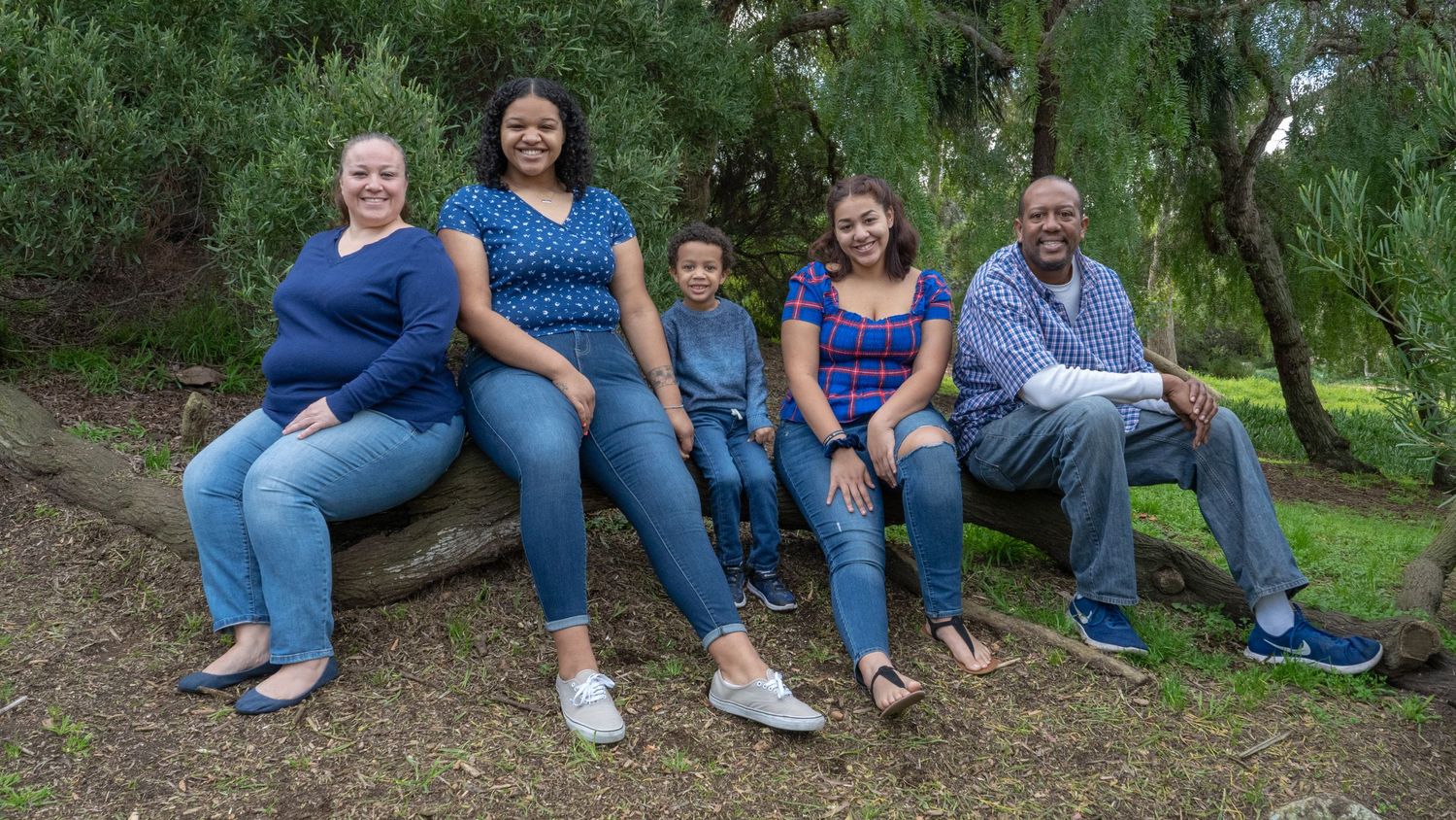Smiling family of five sitting casually on a log outdoors surrounded by green trees.
