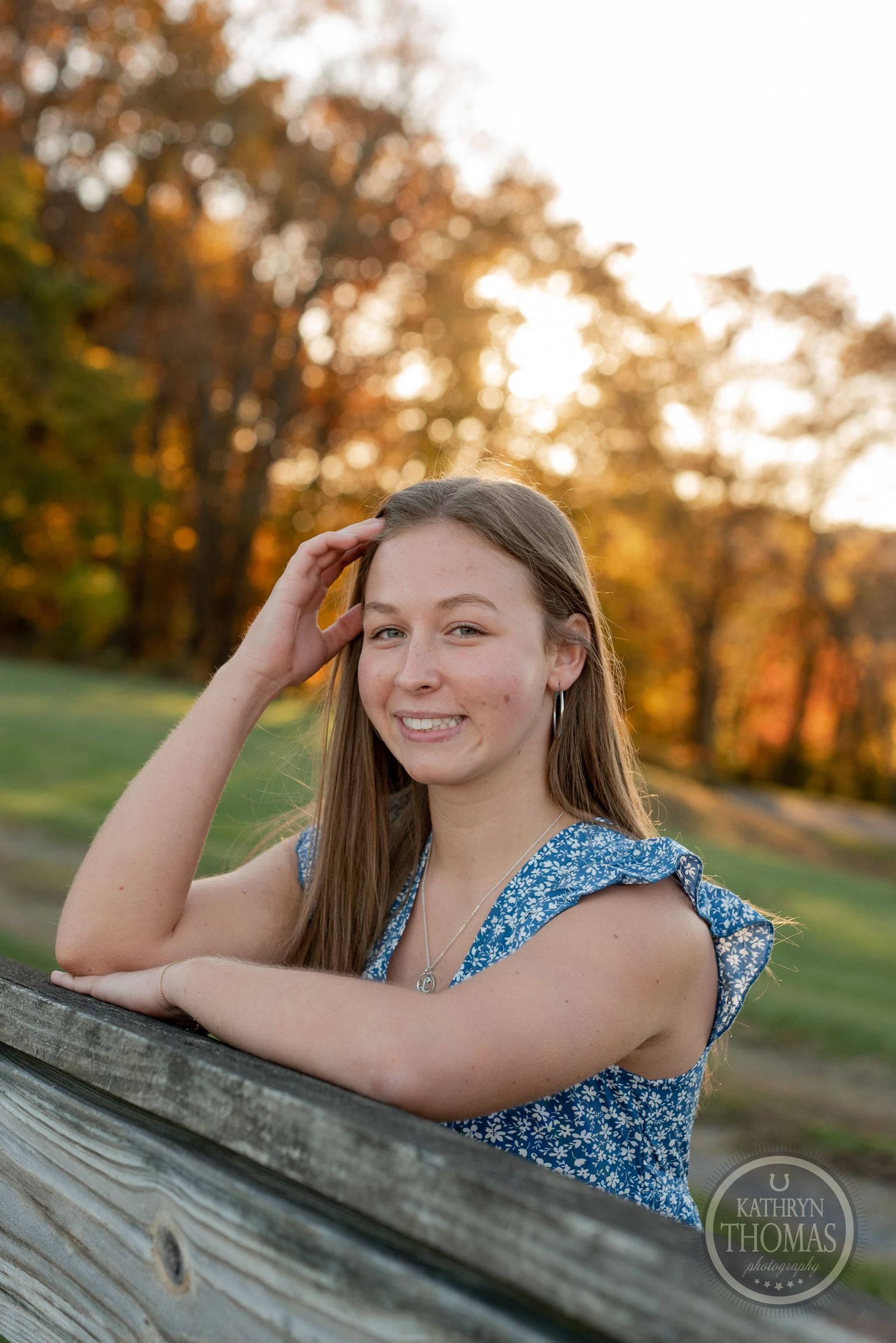 Someone in a sleeveless blue dress smiles while leaning on a wooden fence at sunset.
