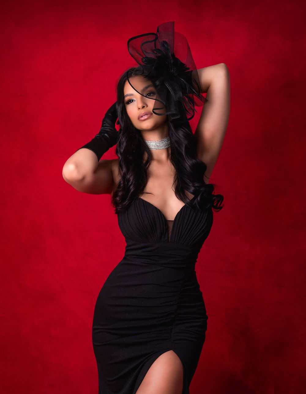 Woman in black evening gown and bow headpiece poses against red backdrop.