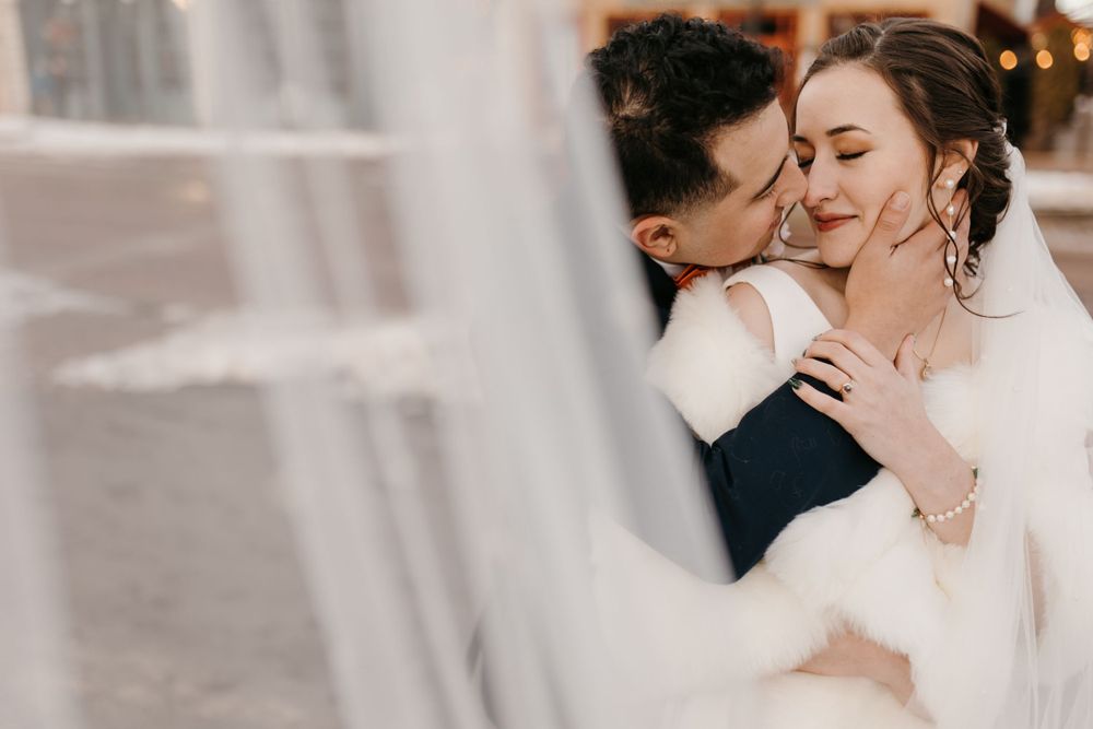 Groom gently kisses bride's cheek as she smiles, both embracing warmly in a close winter wedding portrait outdoors.
