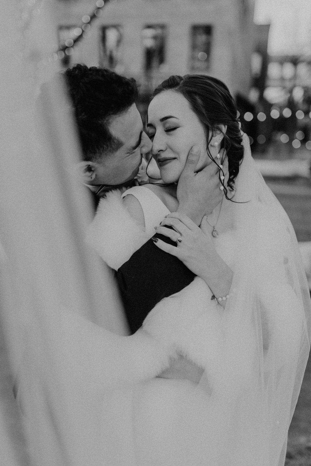 Black and white romantic portrait of groom kissing bride's cheek as she closes her eyes, veil framing the couple.