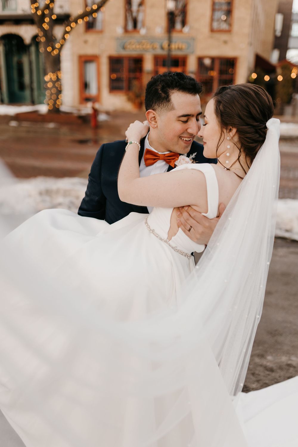 Smiling groom in orange bow tie lifts bride in flowing white dress outside festive winter venue.
