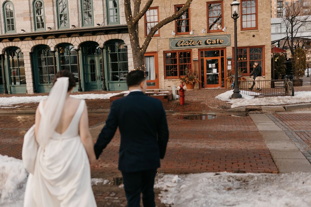 Bride and groom walk hand in hand toward a brick building decorated with string lights in winter.