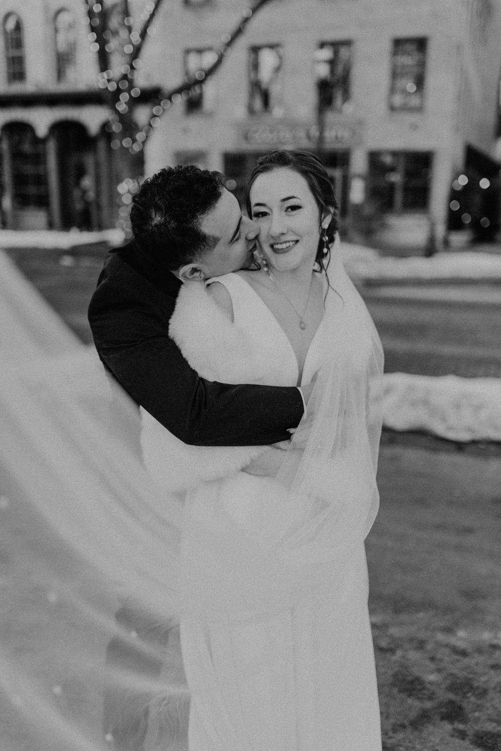 Black and white photo of groom kissing bride's cheek as she smiles, with flowing veil and bokeh lights behind them.