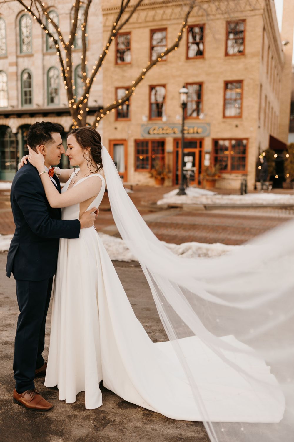Bride and groom embrace nose-to-nose in a snowy town square, bride's cathedral veil flowing dramatically in the wind.