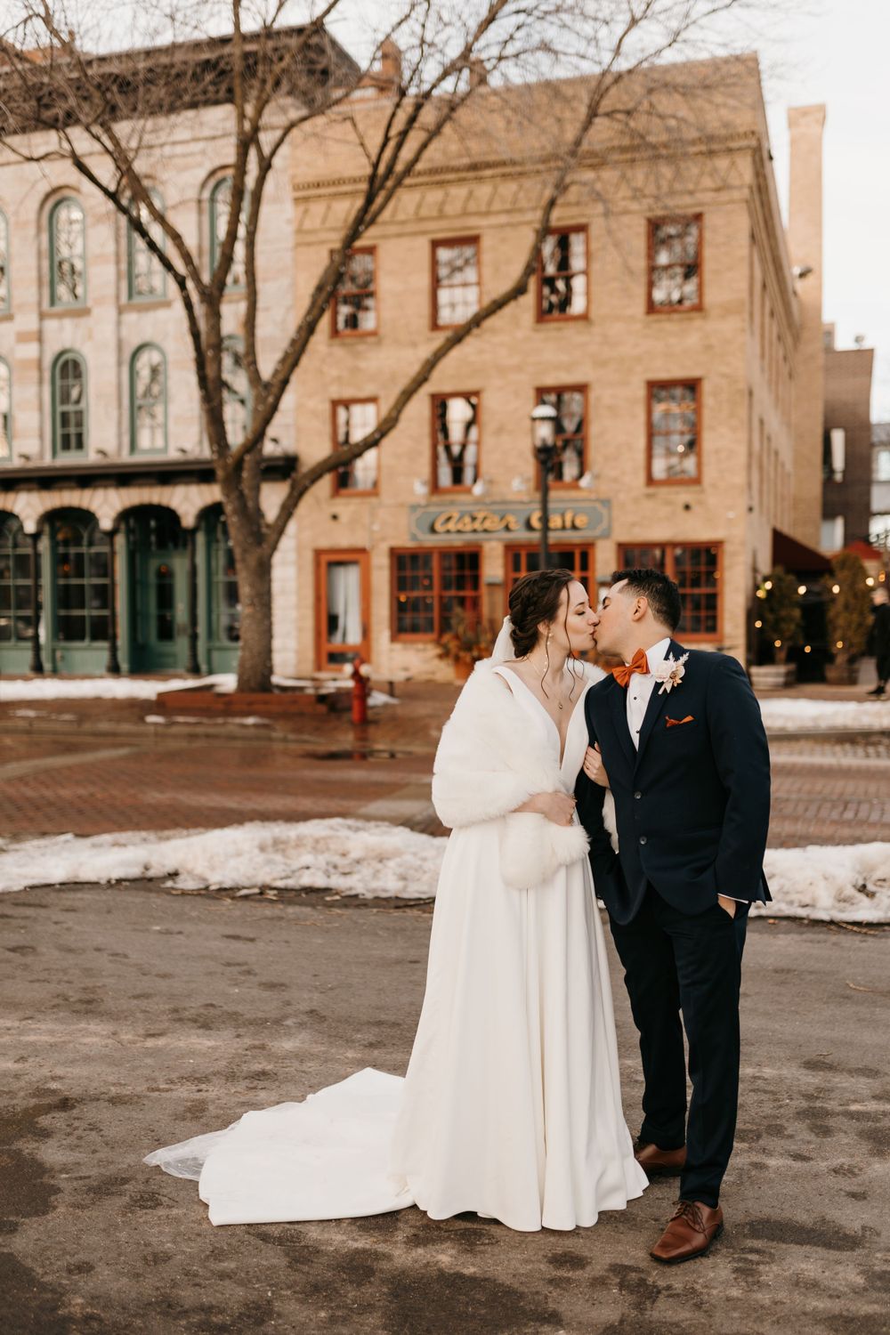 Bride and groom share a kiss in a snowy winter street, bride wearing white gown with fur wrap and flowing train.