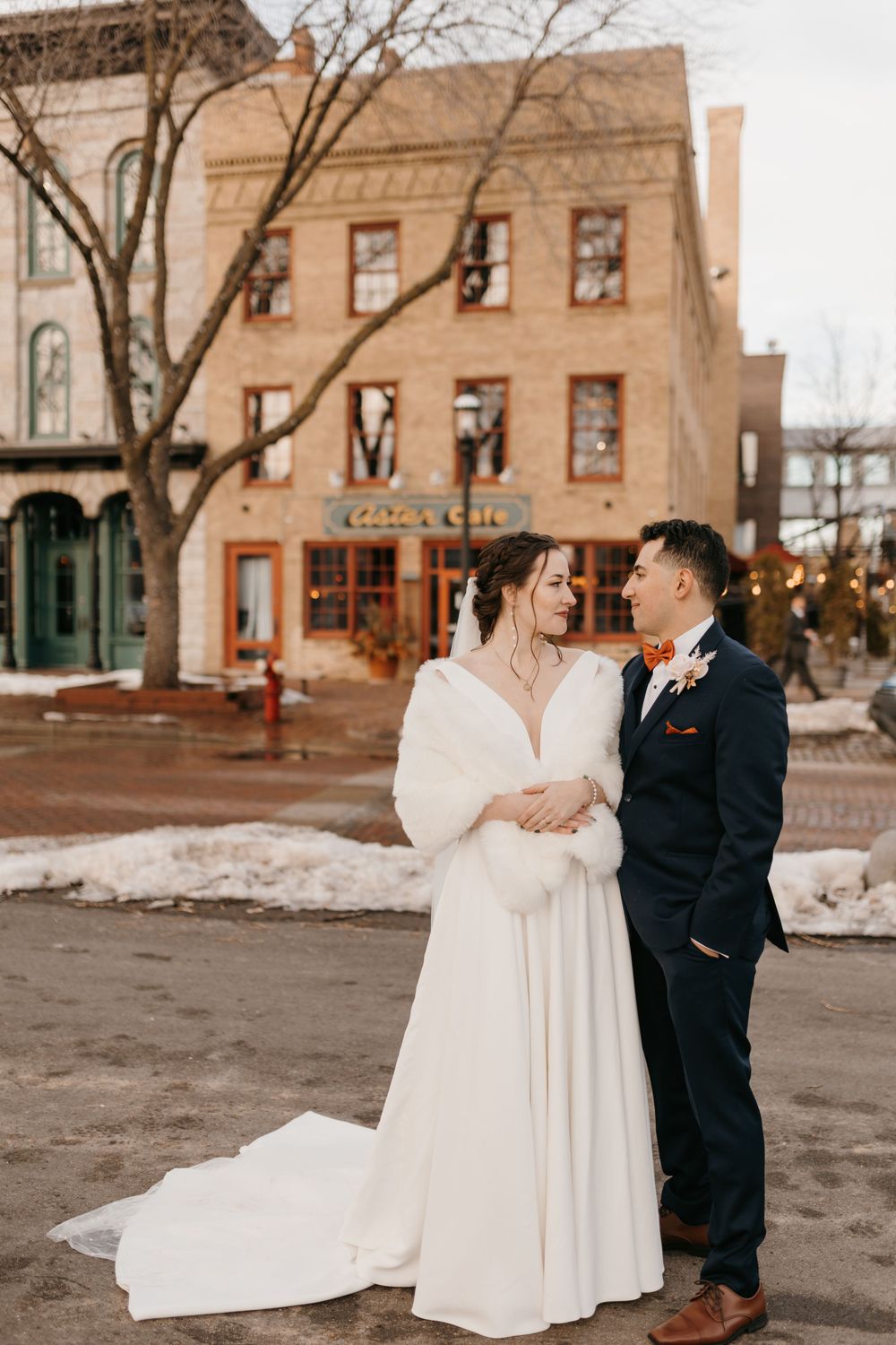Bride and groom stand together in snowy town square with historic brick buildings behind them.