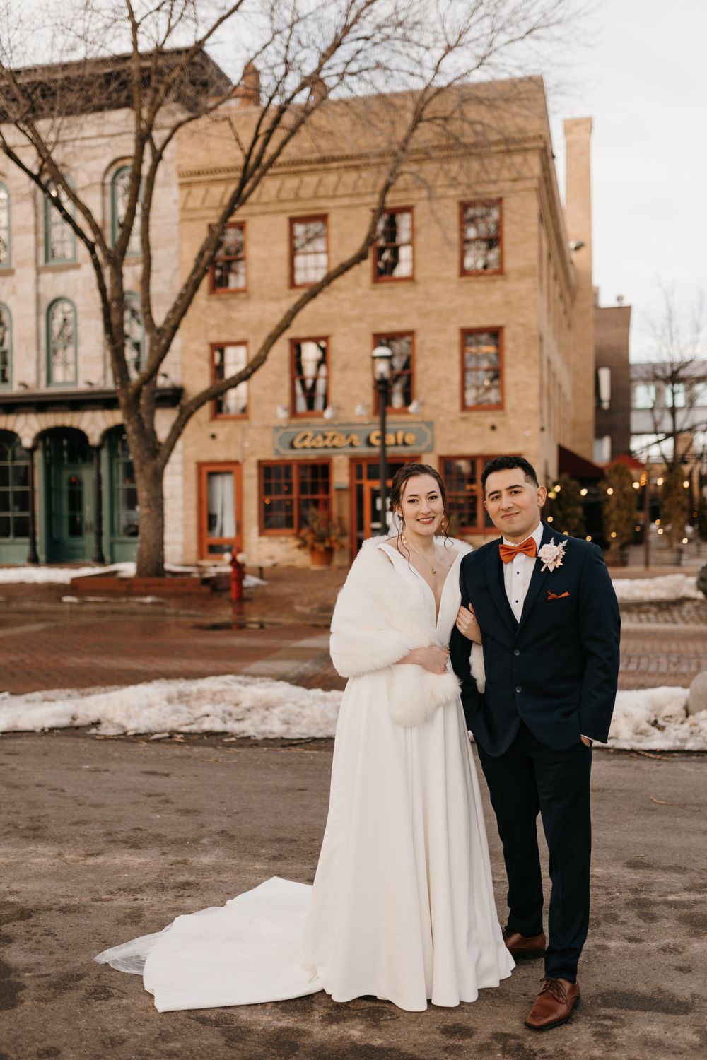 Smiling bride and groom pose together in snowy square with historic Aster Cafe building behind.