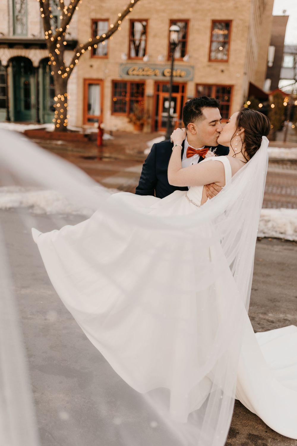 Bride and groom share a kiss as her white veil billows in the wind on a snowy city street.