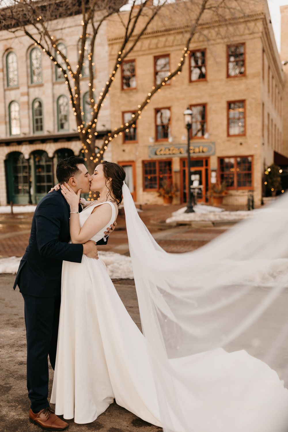 Bride and groom share a romantic kiss outdoors in winter, bride's long white veil billowing in the breeze behind them.