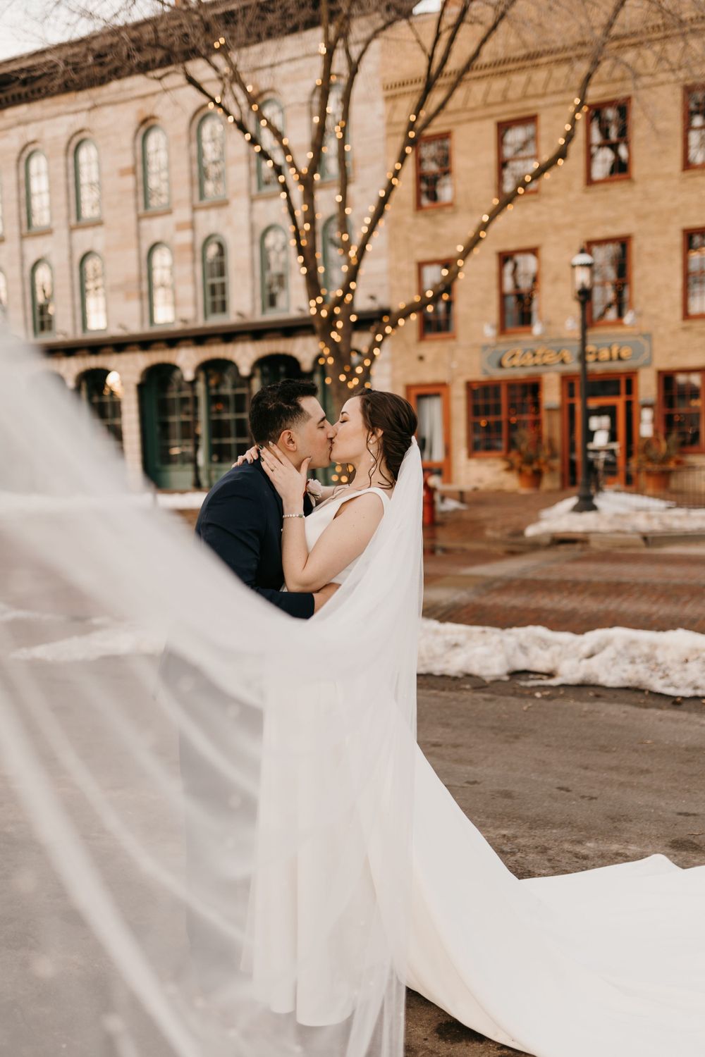 Couple kisses in a charming snowy town square with string lights, bride's white veil sweeping across the foreground.