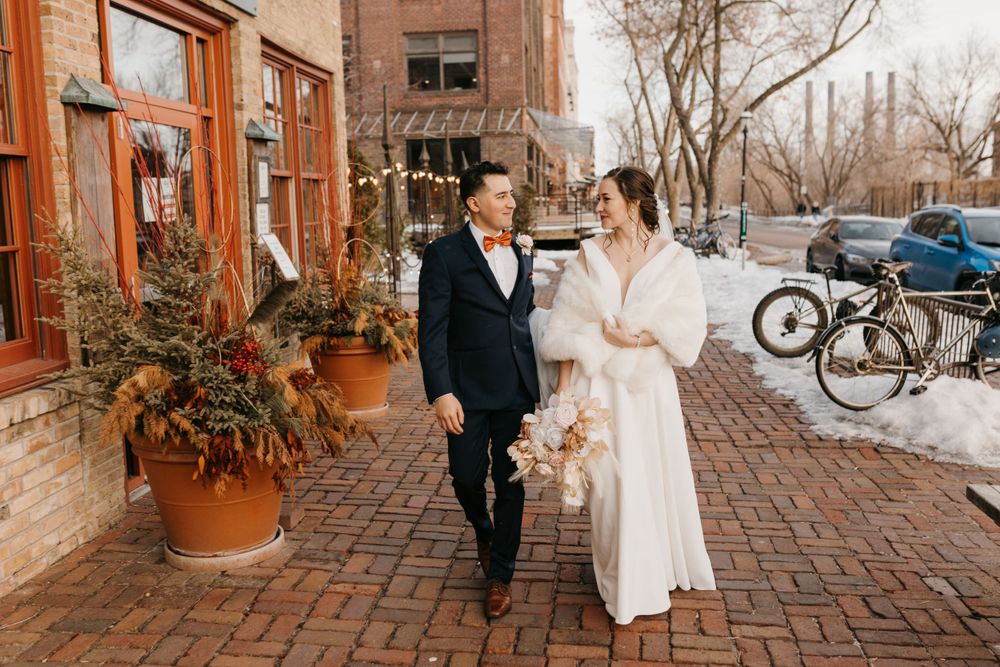 Bride and groom walk together on snowy brick street past festive potted plants in winter.
