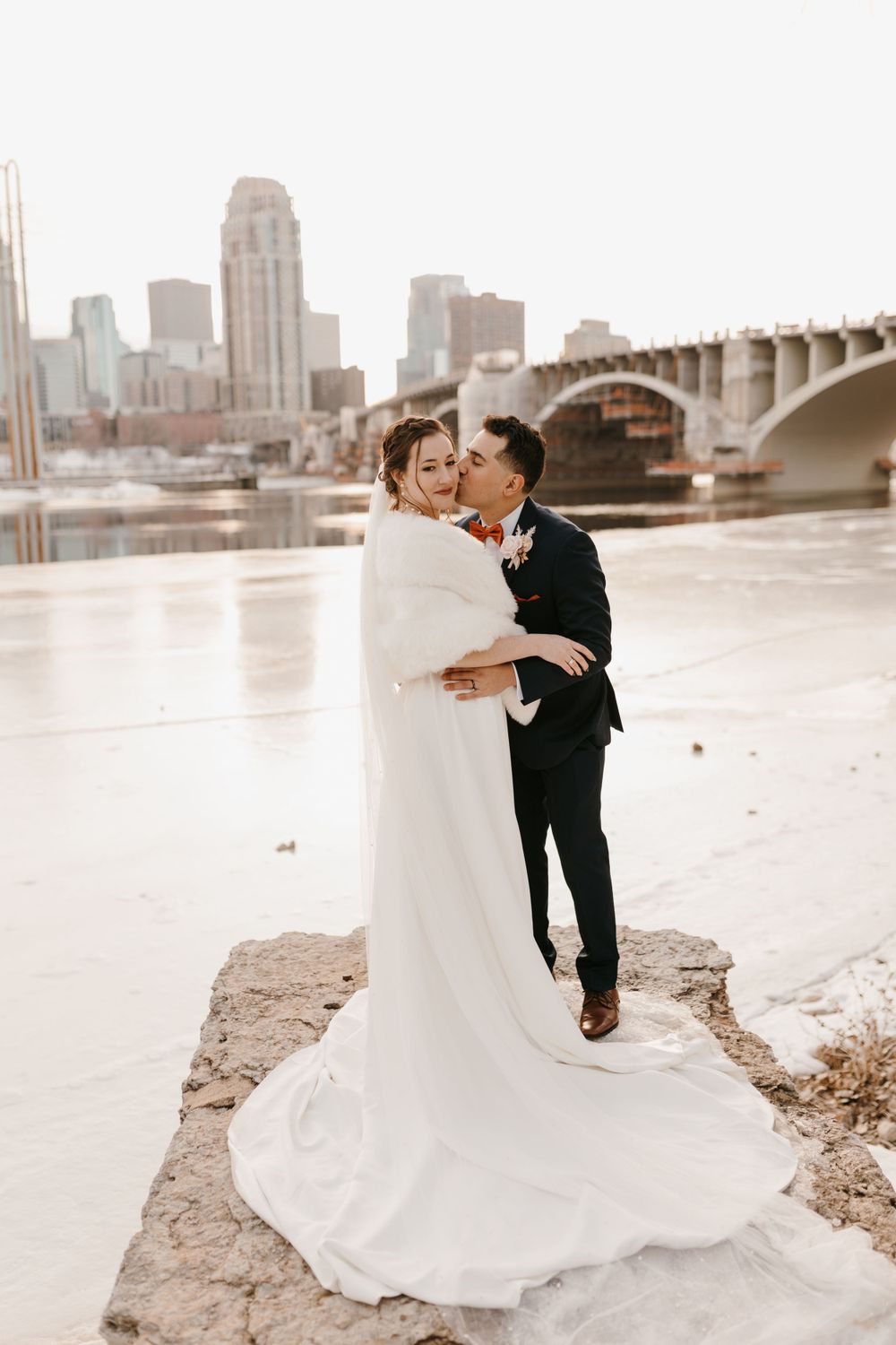 Groom kisses bride on snowy riverbank with city skyline and bridge visible in background.