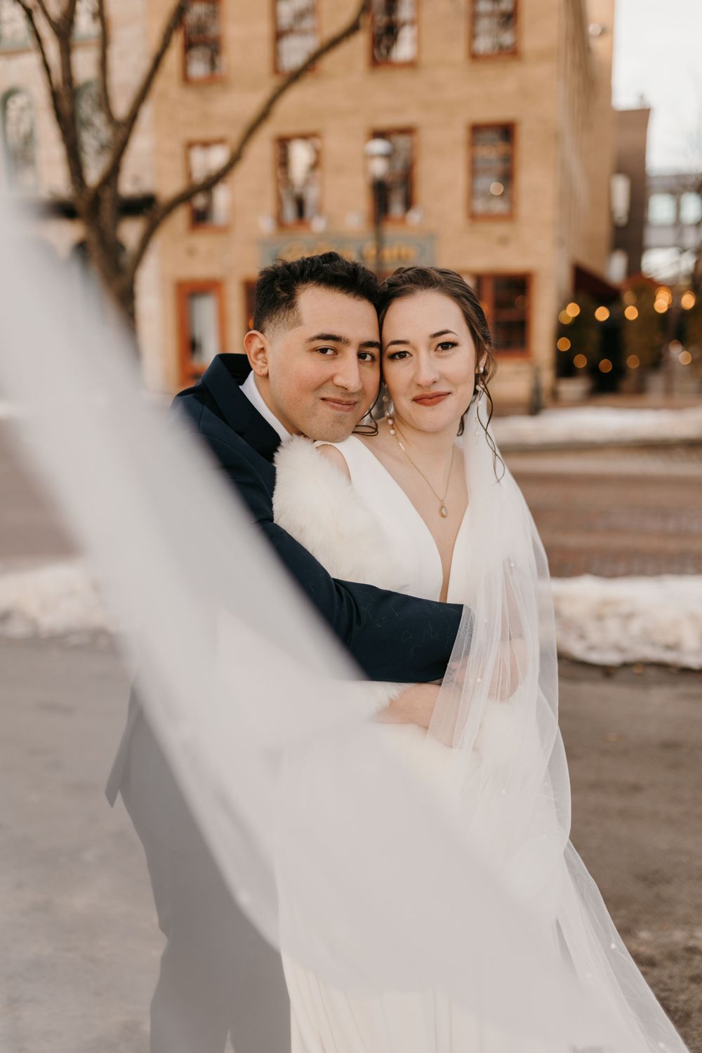 Groom embraces bride from behind outdoors in winter, her veil flowing in the foreground of the romantic portrait.