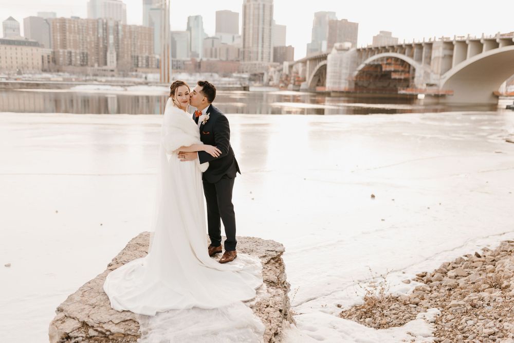 Wedding couple poses on snowy riverbank rocks with Minneapolis skyline and bridge behind them.