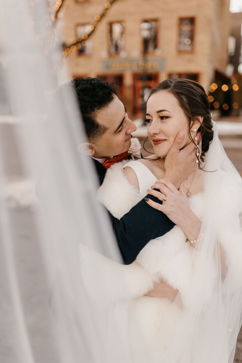 Groom tenderly cups bride's face while leaning in close, bride smiling softly in a white gown and fur wrap outdoors.