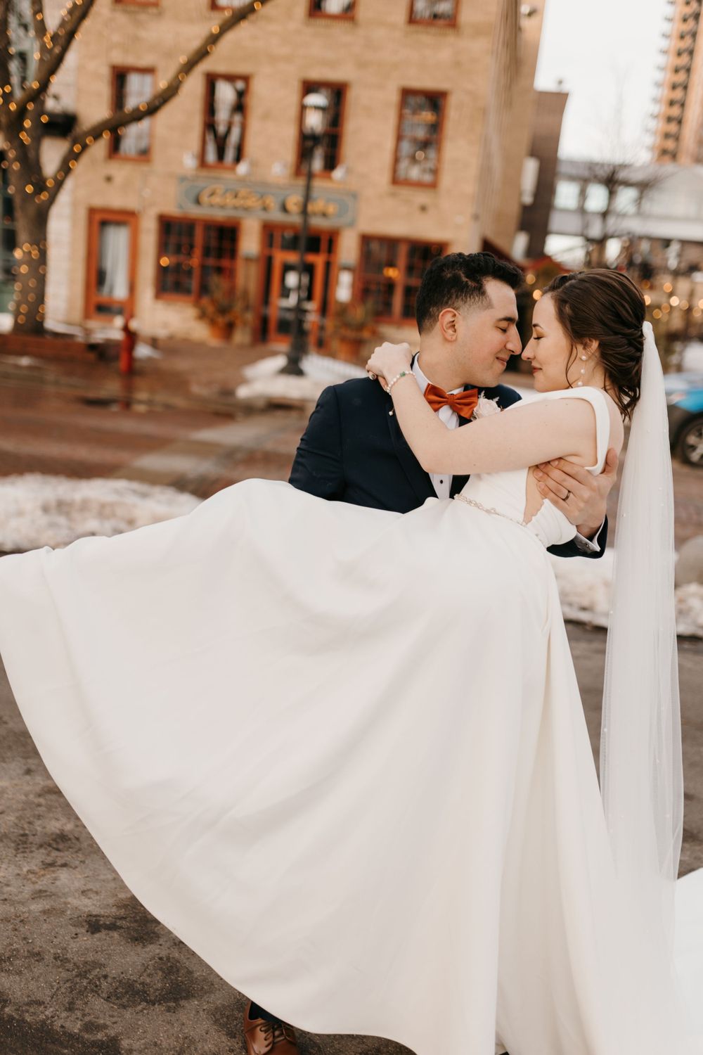 Groom in navy suit lifts bride in white gown and veil, sharing romantic moment on snowy winter street.
