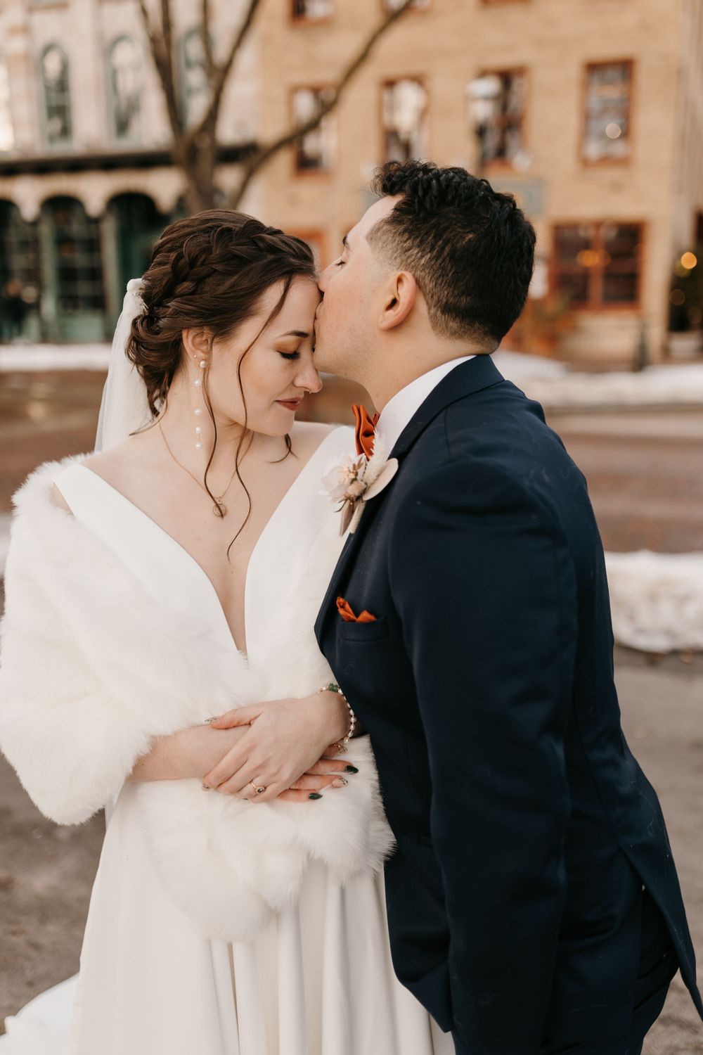 Groom tenderly kisses brides temple outdoors in winter, bride wearing fur wrap and veil.
