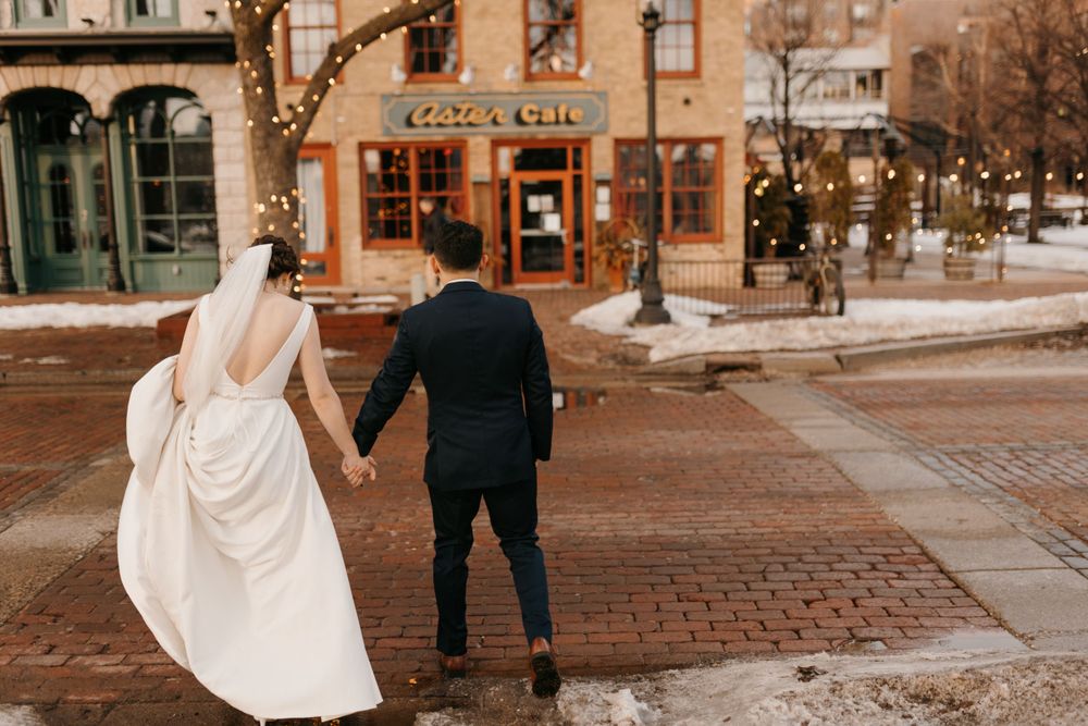 Wedding couple holds hands walking across brick plaza toward a cozy cafe with warm string lights.