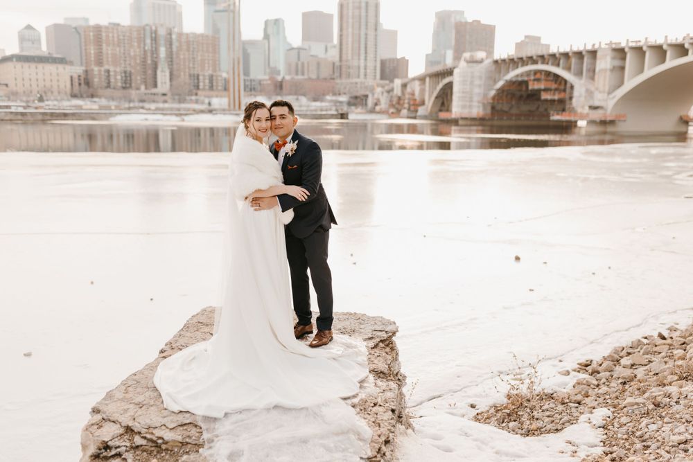 Bride and groom embrace on rocky riverbank with city skyline and bridge in winter background.
