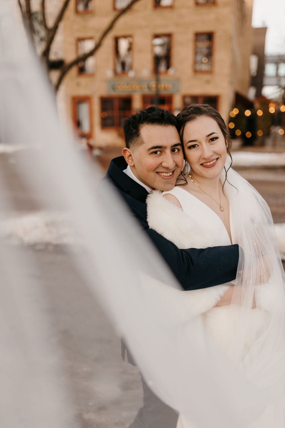 Smiling bride and groom pose closely together in a winter outdoor setting with bride's veil dramatically in foreground.