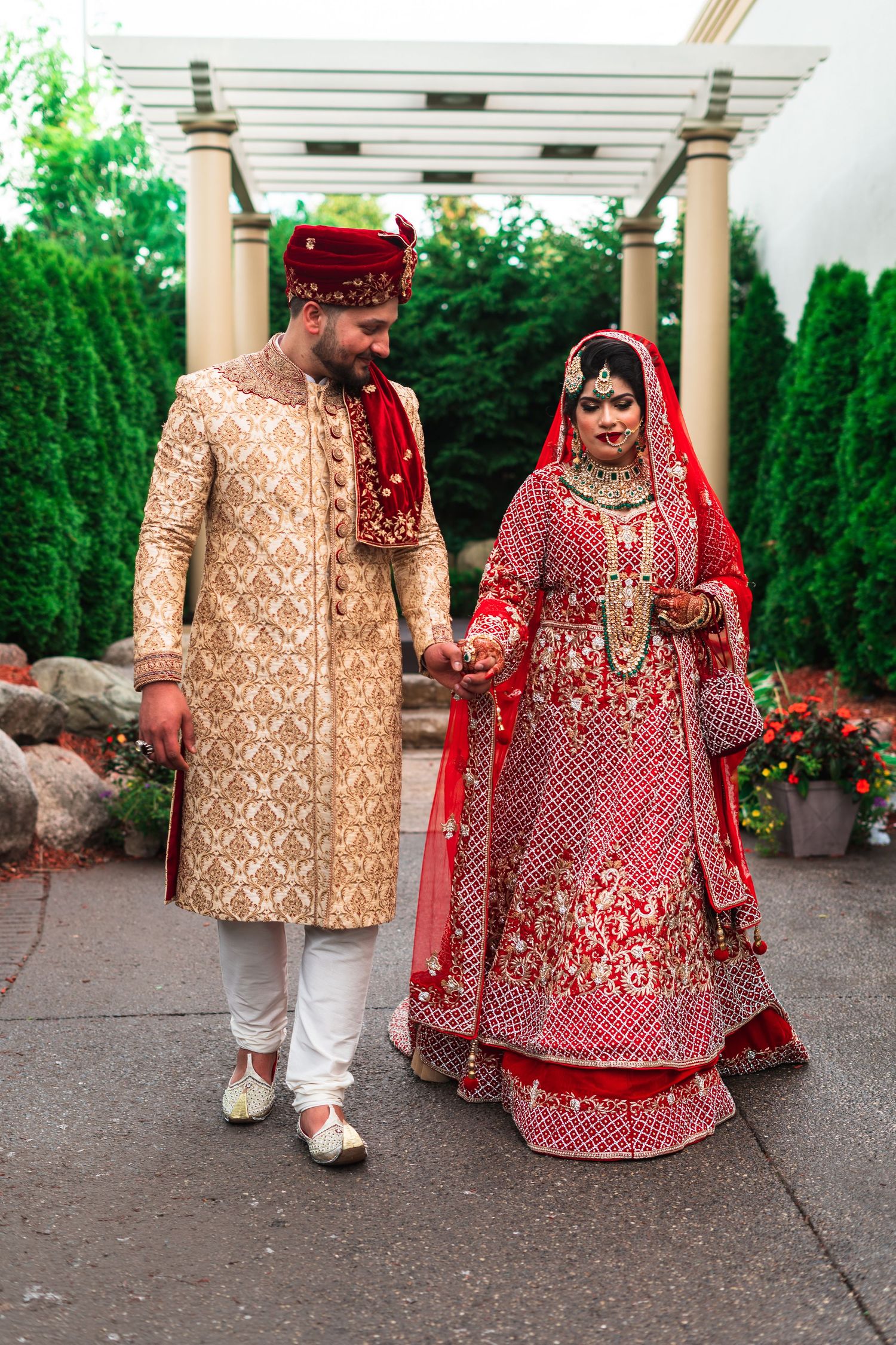 Traditional Indian wedding couple in ornate red and gold attire standing under garden pergola.