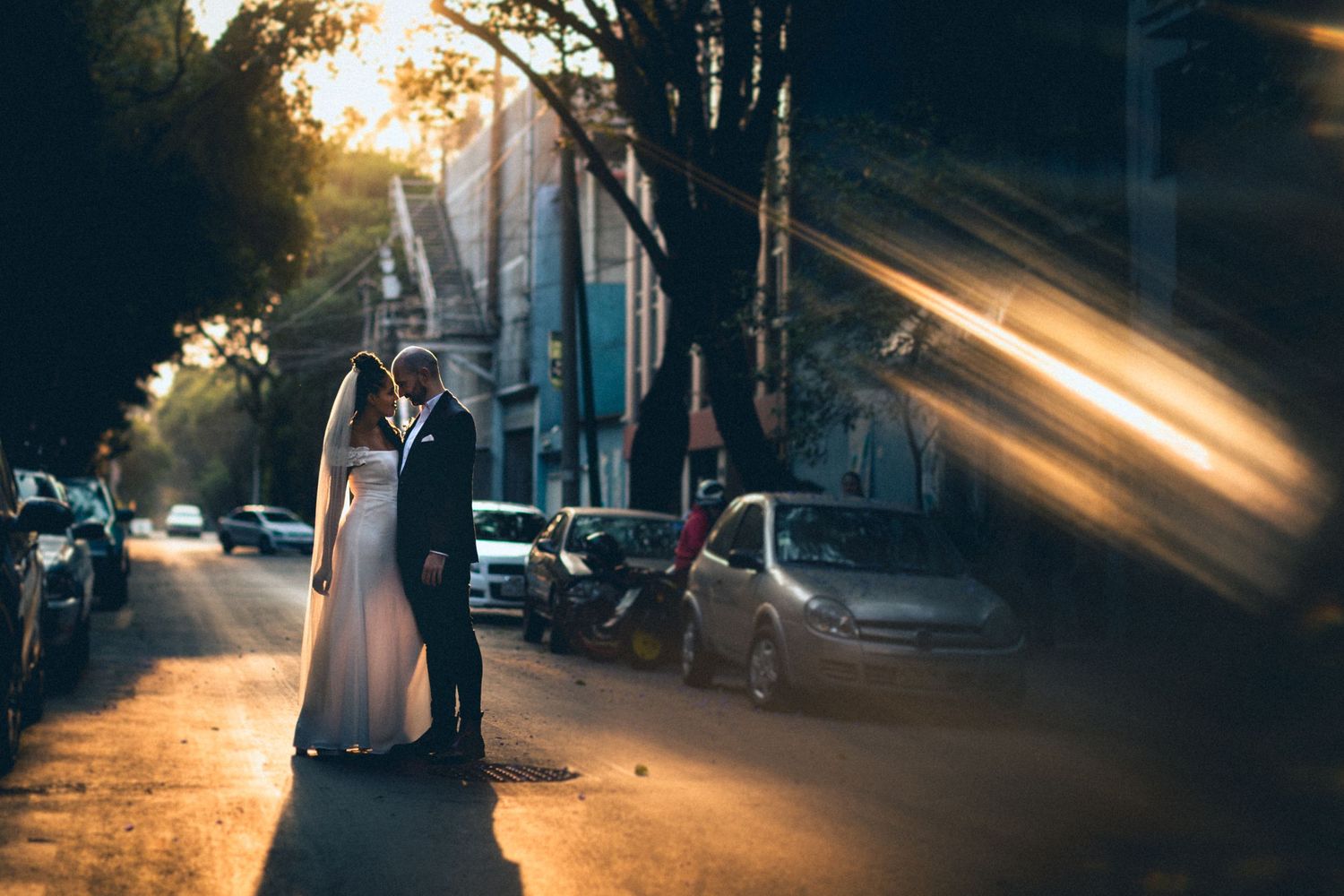 A wedding couple embraces on a sunset-lit street with dramatic light rays.