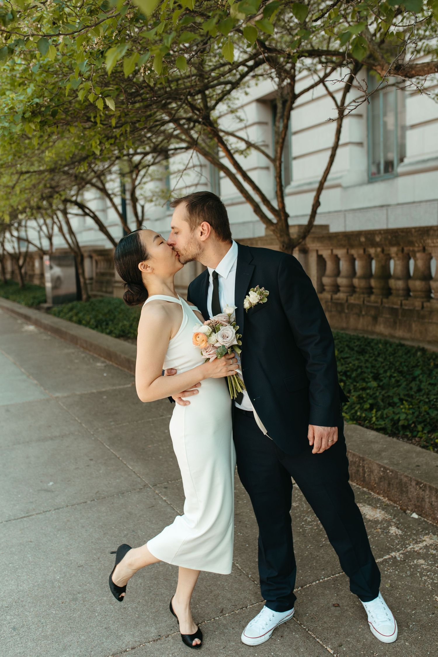 Couple in formal attire shares romantic moment on sidewalk with bouquet.