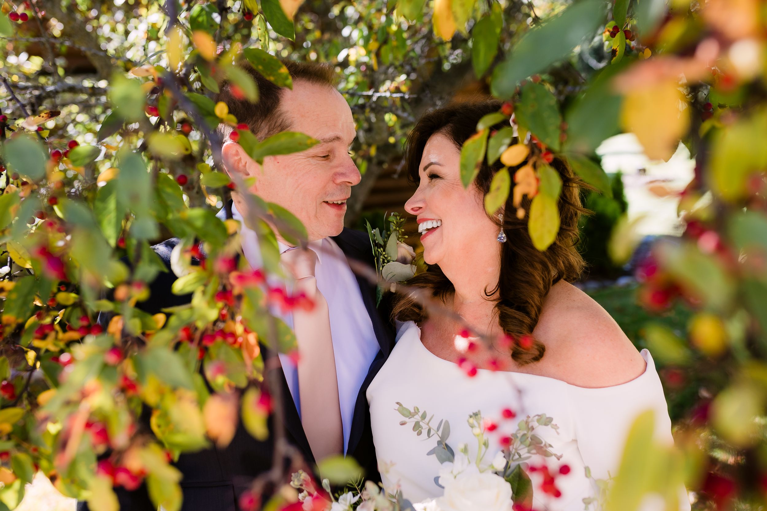 Bride and groom embracing behind a green and red tree. Smiling at each other.