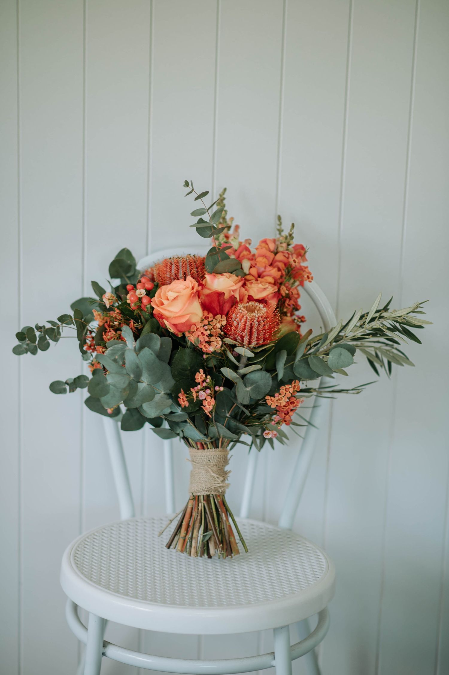 A elegant bridal bouquet with coral roses, eucalyptus, and greenery tied with twine on a white chair.